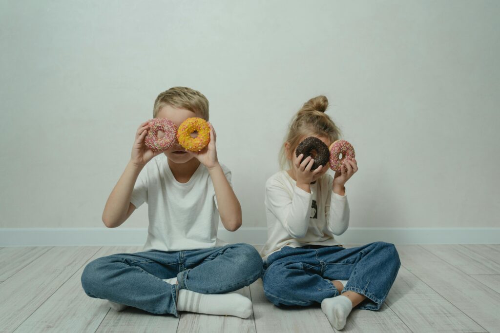 Two children playfully holding donuts over their eyes indoors on a wooden floor.