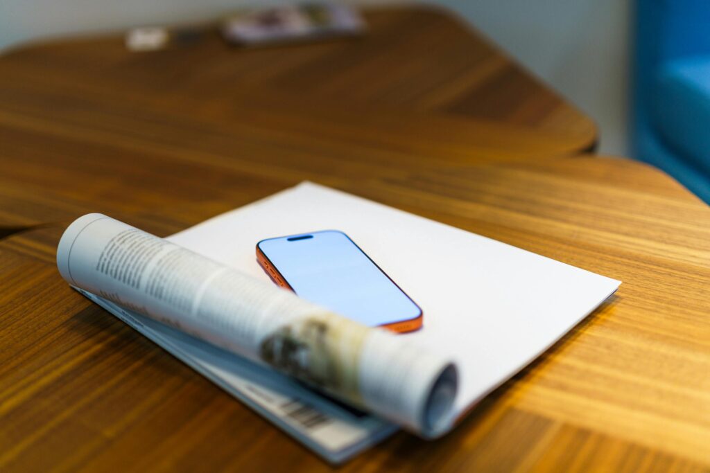 A sleek mobile phone on a wooden table with a magazine and blank paper, offering perfect copyspace.