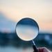 A magnifying glass held by a hand outdoors at sunset, focusing on exploration.
