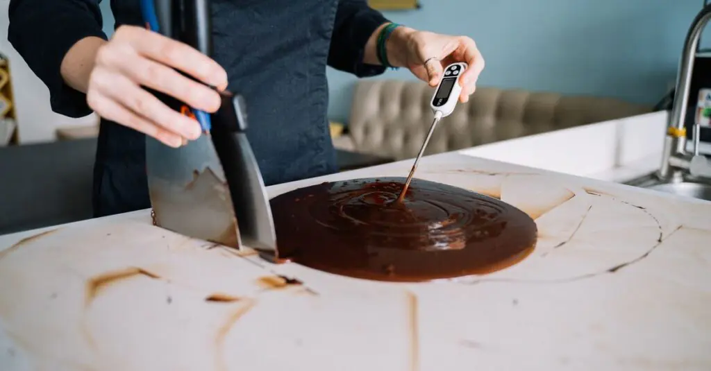 Close-up of a person tempering chocolate on a kitchen counter using a scraper and thermometer.