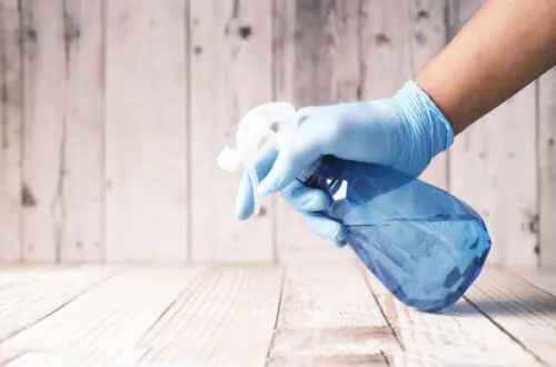 A gloved hand holding a blue spray bottle against a wooden background.