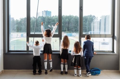 A group of school children in uniform stand by a large window, looking outside.