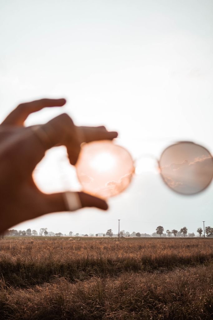 A hand holds sunglasses reflecting a beautiful sunset over a country field, creating a warm, dreamy effect.