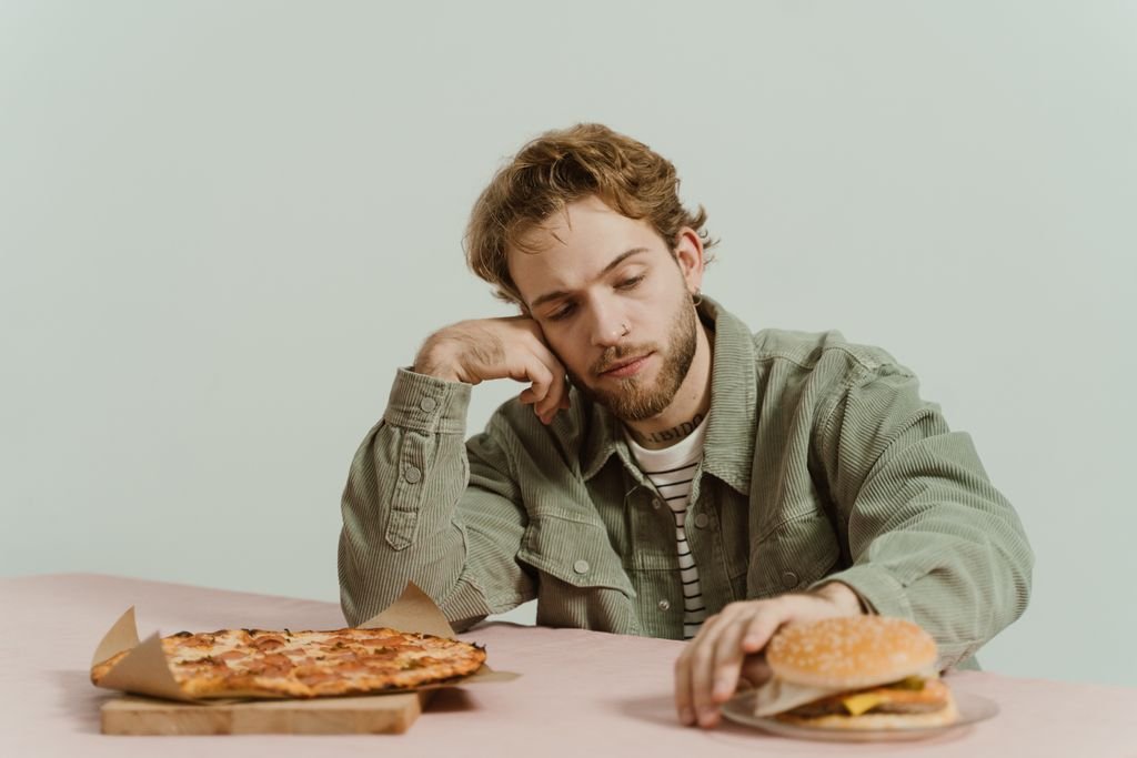 A man sits thoughtfully at a table with a pizza and burger in a minimalist indoor setting.