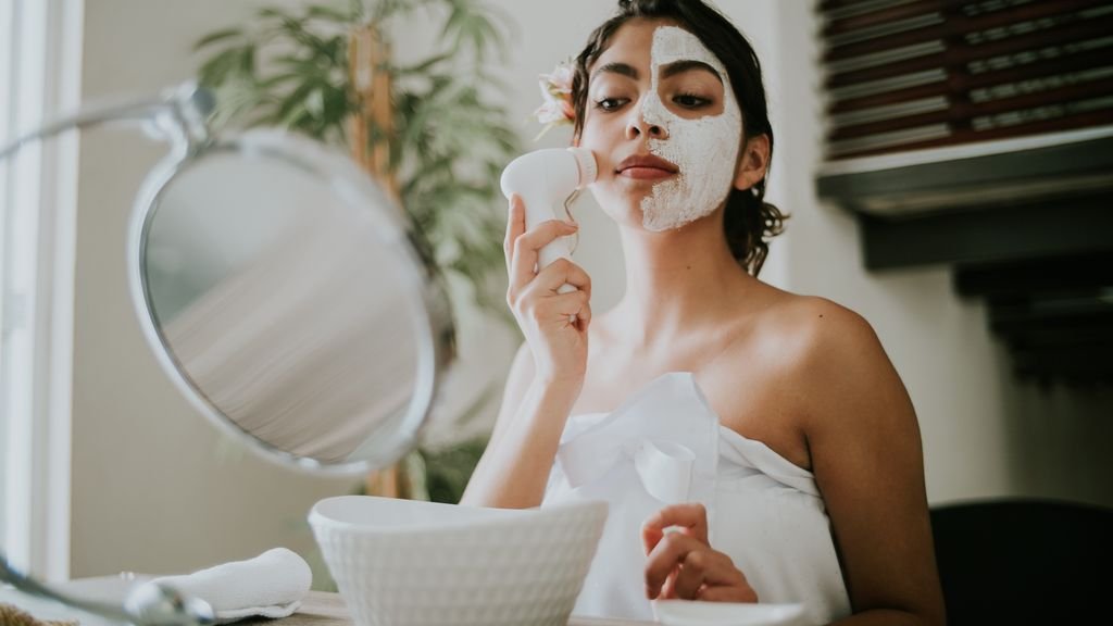 A woman uses a facial brush to apply a clay mask during a relaxing skin care routine.