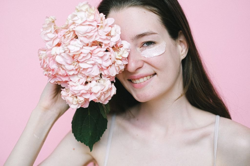 Crop young woman hiding eye behind pink hydrangea while standing with moisturizing product on face skin against pink background