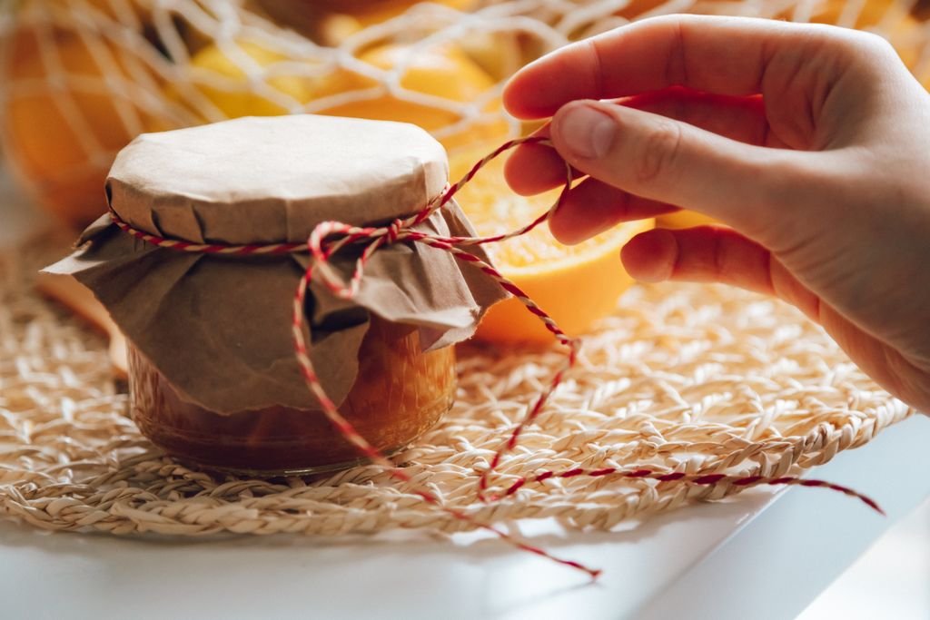 Homemade jam jar with hand tying string, surrounded by fresh oranges in warm lighting. food