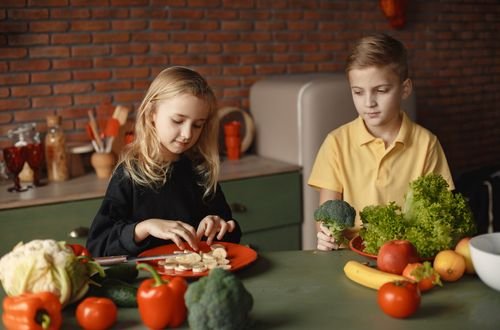 Positive little girl and boy sitting at desk with various fresh vegetables and fruits while preparing meal in loft kitchen with brick wall