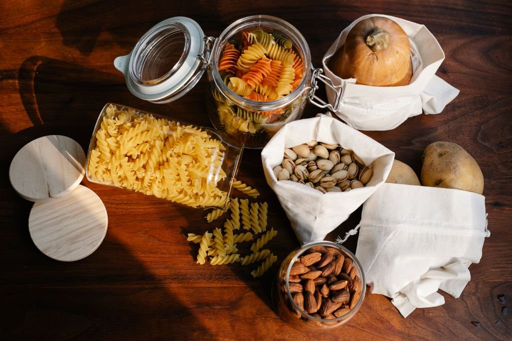 Top view jars of raw pasta placed on wooden table near ECO friendly sacks with pistachios and almonds near pumpkin and potatoes food