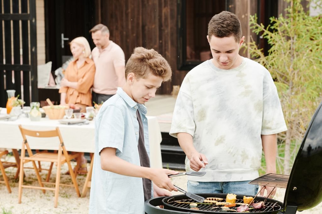 Two boys grilling food at a family barbecue with parents in the background.