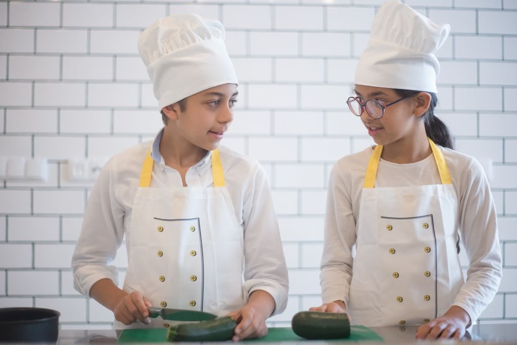 Two children in chef attire preparing vegetables in an indoor kitchen setting.
