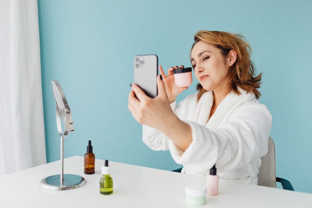 Woman in bathrobe taking a skin care selfie with products on desk.