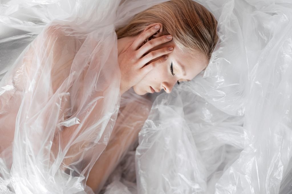A peaceful woman with closed eyes lies wrapped in translucent plastic, symbolizing fragility and beauty. nylon